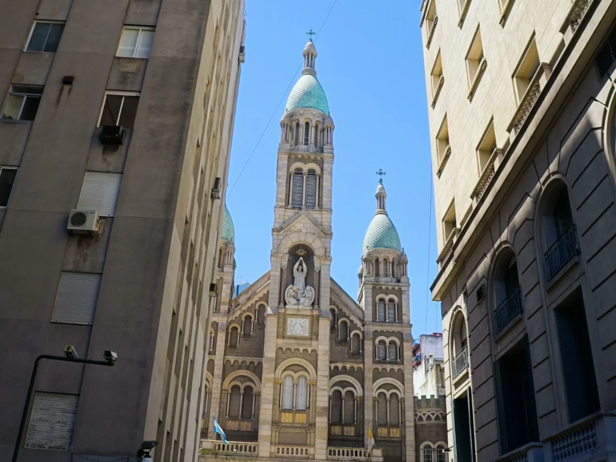 Basilica of the Blessed Sacrament A grand basilica with green domes and intricate stonework, framed by two modern city buildings under a clear blue sky.