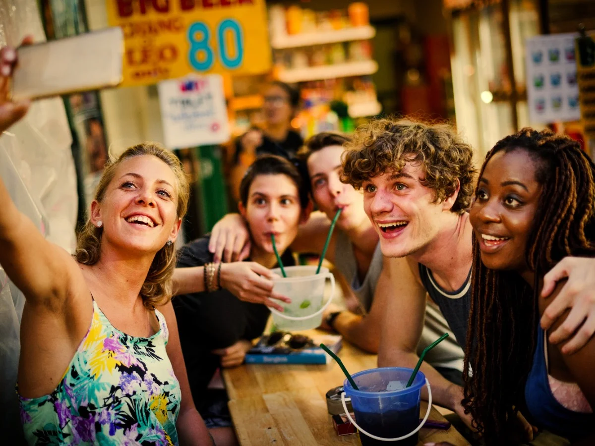 Friends Taking Selfie with Drinks A diverse group of five young adults happily taking a selfie while drinking beverages with straws from buckets, likely at a casual outdoor eatery or market.
