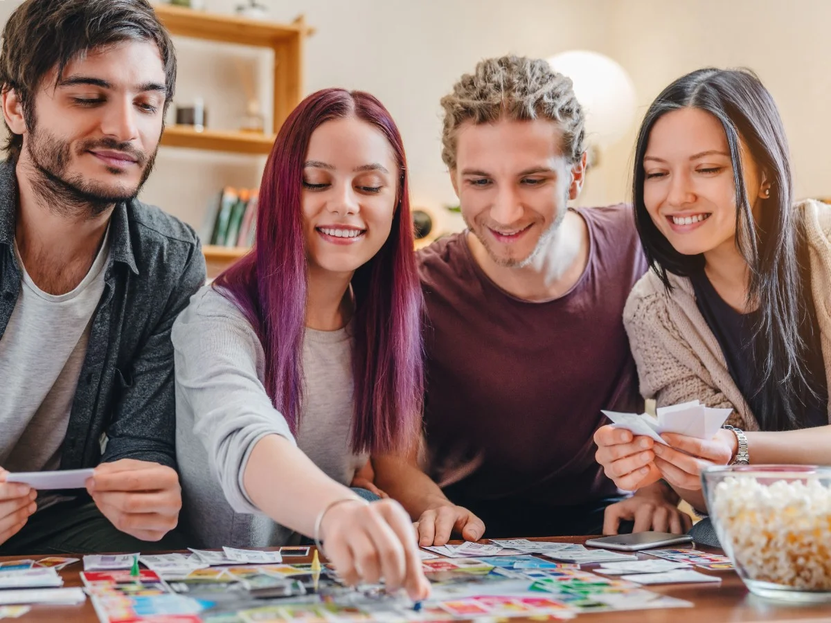 Team Playing Board Game Four smiling young adults gathered around a table, engaged in a board game with cards and game pieces, a bowl of popcorn visible.
